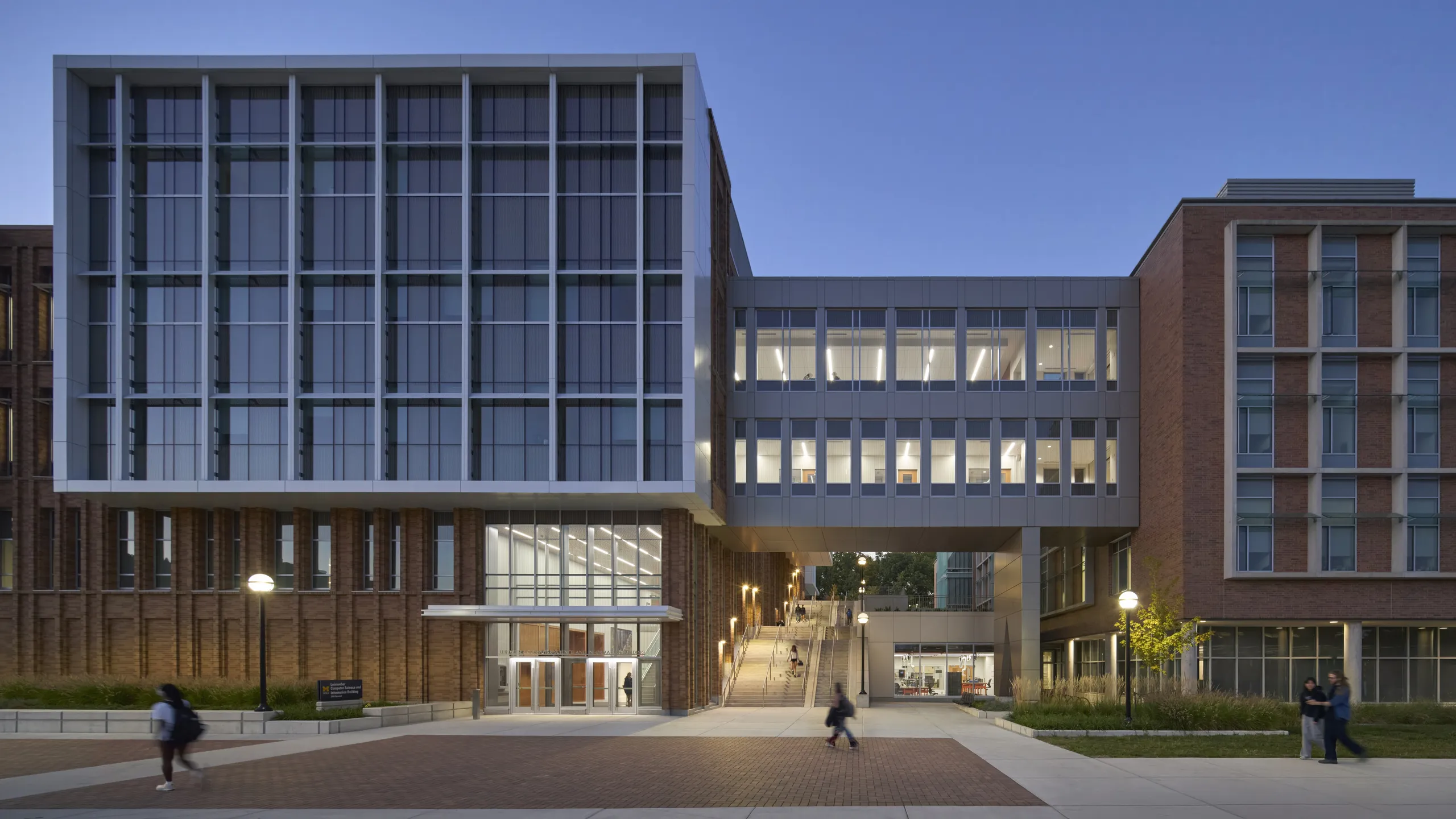 Modern glass-and-brick university building in Michigan, illuminated at dusk with students walking by.